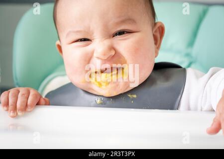 Ein dreckiges, fröhliches Baby am Tisch auf einem Stuhl hat seinen Mund mit Gemüsepüree schmutzig gemacht. Einführung ergänzender Lebensmittel Stockfoto