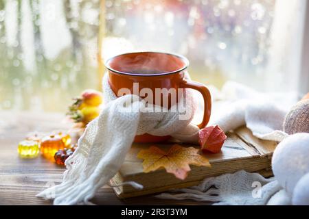 Ein Orangenbecher in einem Schal mit heißem Tee, Kürbissen, gelben, trockenen Ahornblättern, ein Buch auf dem Fensterbrett, Regentropfen am Fenster - au Stockfoto