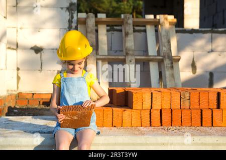 Ein kleines Mädchen mit gelbem Schutzhelm spielt Baumeister auf der Baustelle ihres zukünftigen Hauses. Erwartung, sich zu bewegen, zu wählen Stockfoto