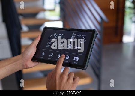 Hände eines birassischen Mannes zu Hause, der ein Tablet mit Smart-Home-Schnittstelle auf dem Bildschirm verwendet Stockfoto
