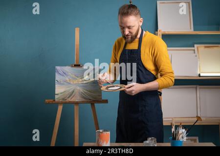 Ein bärtiger männlicher Künstler in einer schwarzen Schürze steht in einem Kunststudio mit einer Palette mit Farben in den Händen und schaut auf den leeren Platz für die Stockfoto