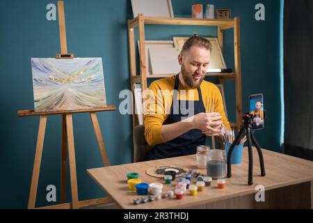 Kunstschule. Malerblog. Glücklicher Maler am Tisch mit künstlerischen Werkzeugen, der im Kunstatelier auf ein Smartphone schaut. Online-Kurse Stockfoto