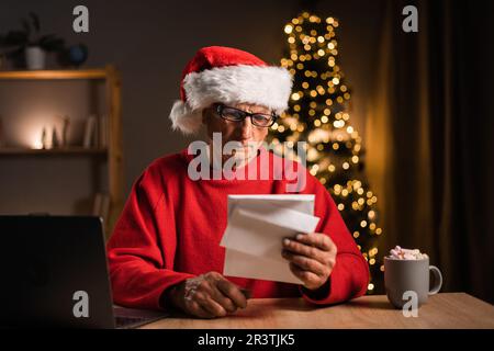 Der weihnachtsmann sitzt am Tisch im Nordpolhaus und liest Weihnachten Weihnachten Wunschzettel-beeindruckendes Geschenk mit Neujahrsdekoration drinnen Stockfoto
