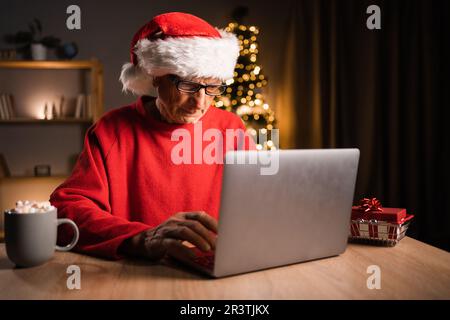 Lächelnder älterer Mann mit Weihnachtsmann-Hut, der am Heiligabend zu Hause E-Mails auf einem Laptop versendet. Stockfoto