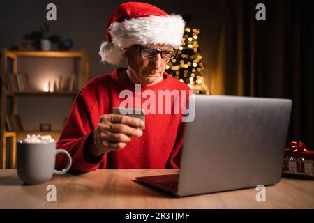 Weihnachtseinkäufe Online. Seniorenmännchen mit weihnachtsmannmütze mit Kreditkarte und Laptop, der zu Hause im Wohnzimmer sitzt und Geschenke bestellt. Der Weihnachtsmann Stockfoto