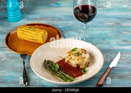 Fleischschnitzel unter Käse mit Spargel und gegrilltem Mais auf blauem Hintergrund Stockfoto