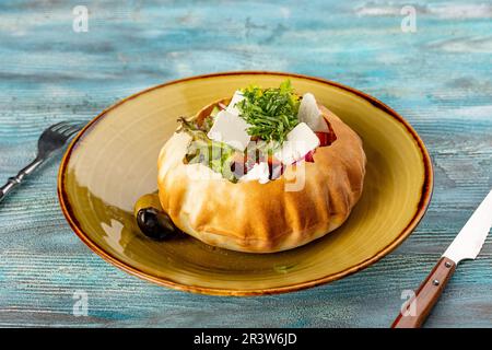 Frischer Gemüsesalat mit Feta im Brot - gesundes Essen. Original-Restaurant, serviert in Brot statt auf einem Teller mit blauem Holzhintergrund Stockfoto