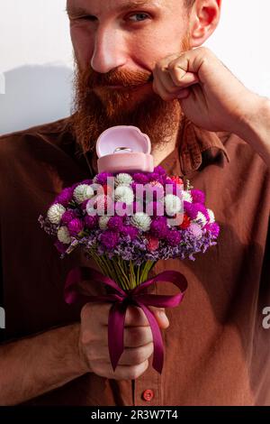 Heiratsantrag. Ein hübscher, eleganter Mann im braunen Hemd gibt einen weißen goldenen Ring in einer pinkfarbenen Schachtel, wunderschöne Blumen, Blumenstrauß. Ein bärtiger Kerl, der Freundin einen Antrag macht Stockfoto