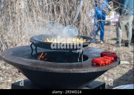 Gemüse wird auf einem runden Herd geröstet. Runder Grill in Form einer Schüssel mit Feuer im Inneren. Mann bereitet Herd für Barbecue zu. Das Brennholz brennt Stockfoto