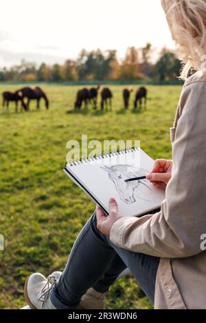 Bleistiftzeichnung. Eine Künstlerin, die ein Porträt eines Pferdes auf der Weide skizziert. Freizeitaktivitäten im Freien Stockfoto