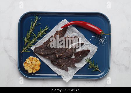 Stücke köstliches Rinderjerky und verschiedene Gewürze auf einem weißen Marmortisch, Draufsicht Stockfoto