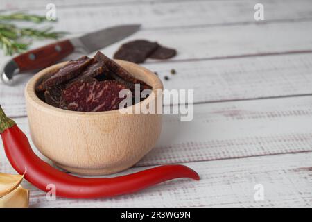 Scheiben von köstlichem Rinderjerky und Chili auf weißem Holztisch, Nahaufnahme. Platz für Text Stockfoto