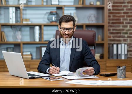 Ein erfahrener Geschäftsmann, der Papierkram erledigt, ein Mann, der mit Dokumenten, Verträgen und Rechnungen arbeitet, sitzt am Tisch mit einem Laptop bei der Arbeit, ein Buchhalter mit Bart und Brille. Stockfoto