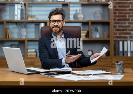 Porträt eines erwachsenen Geschäftsmanns am Arbeitsplatz, eines Mannes mit lächelnden Dokumenten, der vor die Kamera schaut, des Papierkrams eines erfahrenen Chefs am Schreibtisch. Stockfoto