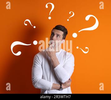 Berufswahl oder andere Lebensbereiche, Konzept. Entscheidungen treffen, aufmerksamer junger Mann, umgeben von gezeichneten Fragezeichen auf orangefarbenem Hintergrund Stockfoto