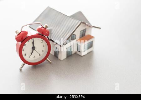 Wecker und Hausmodell. Zeit für eine Hypotheken- oder Immobilienversicherung Stockfoto