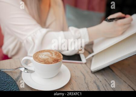 Zugeschnittenes Bild eines Mädchens, das sich beim Kaffeetrinken Notizen in einem offenen Notizblock macht Stockfoto