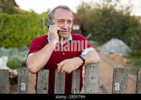 Ein erwachsener Mann, der entspannt neben dem Zaun steht und im Garten telefoniert und lächelt Stockfoto