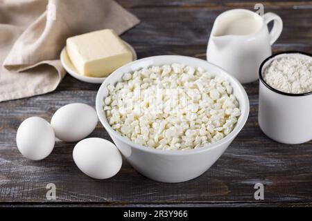 Schüssel mit Hüttenkäse, Eiern, Mehl, Butter, Milch auf Holzhintergrund Stockfoto