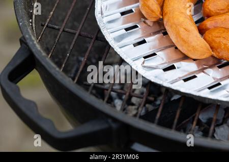 Frische Würstchen auf einem Grillgitter im Garten. Würstchen auf einer Aluminiumschale. Foto bei natürlichen Lichtverhältnissen. Stockfoto