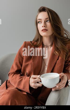 Brünette junge Frau mit langen Haaren und Halskette, die Terrakotta und einen trendigen Anzug mit Blazer und Hose trägt und eine Tasse Kaffee mit Schaum hält, während sie si Stockfoto