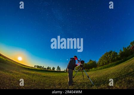 Astronomen, die ein Teleskop im Garten benutzen, in einer hellen Mondnacht, mit dem schwindenden Gibbuus-Mond, der links aufgeht. Stockfoto