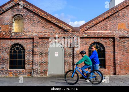 Sandnes, Rogaland, Norwegen, 18 2023. Mai, Mutter geht mit ihrem Sohn auf einem Fahrrad, vorbei An einem traditionellen Ziegelgebäude unter blauem Himmel am Abend Stockfoto