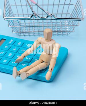 Eine Holzpuppe und ein blauer Taschenrechner, hinter einem Miniatur-Einkaufskorb Stockfoto