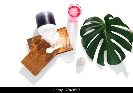 Modell eines Kunststoffbehälters für Kollagenpulver. Löffel mit Kollagen auf Holzbühnen, Glasbecher und Monsterablage auf dem Hintergrund. Stockfoto