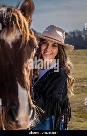 Hübsches braunes Cowgirl, das einen Tag mit ihrem Pferd auf der Farm genießt Stockfoto