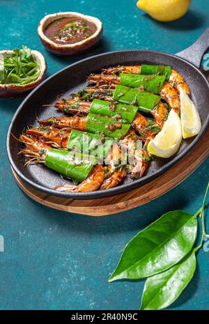 Scharfe Knoblauch-Tigergarnelen Shrimps auf Bratpfanne mit Zitronenblättern auf blauem Bäcker, Nahaufnahme Stockfoto
