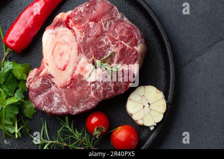 Rohe frische Ossobuco con Kräuter, Knoblauch und Chile Pepper auf schwarzem Teller. Dunkler Hintergrund Stockfoto