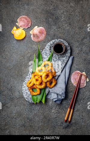 Frittierte Tintenfischringe auf der Steinplatte mit tropischen Blättern verziert. Asiatische Lebensmittel Konzept, Sojasauce und Essstäbchen Stockfoto