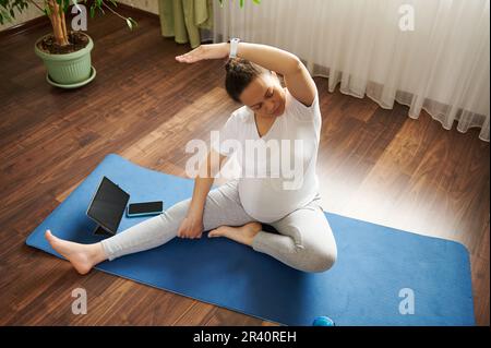 Schwangere Frau übt Online-Schwangerschafts-Yoga auf einer Matte, macht pränatale Dehnung in Körpergleichgewichtsübungen. Draufsicht Stockfoto