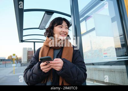 Süßes lächelndes asiatisches Mädchen, das an der Bushaltestelle steht, ein Smartphone in der Hand hält, eine Winterjacke und einen Schal trägt. Frau, die zur Arbeit oder zur Arbeit pendelt Stockfoto