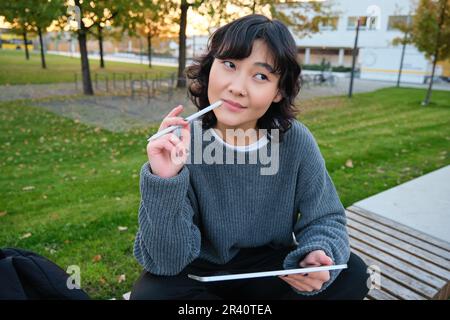 Porträt einer asiatischen Frau, Studentin im Park, sitzt auf einer Bank mit einem digitalen Tablet und einem Stift, denkt, schaut beiseite, umsichtig, geschminkt Stockfoto