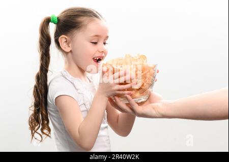 Das kleine Mädchen erhält eine große Schüssel Chips Snacks mit Schmalz, weißes Hintergrundporträt eines kleinen Mädchens, das Chips isst, ein Kind und ungesundes Fast Food. Stockfoto