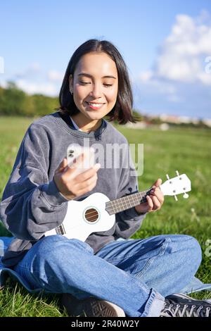 Lächelndes asiatisches Mädchen, das sich die Gitarre-App ansieht, Ukulele singt und spielt, während es auf dem Smartphone in die Akkorde schaut und drinnen im Freien sitzt Stockfoto