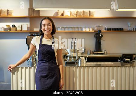 Porträt einer lächelnden asiatischen Barista, die eine Schürze trägt, mit Kaffee an der Theke steht, im Café arbeitet Stockfoto