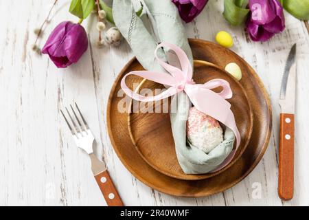 Ostertisch mit bemalten Eiern, lila Tulpe und Besteck auf festlichem Tisch. Tischeinstellung für Happy Easter Day. Stockfoto