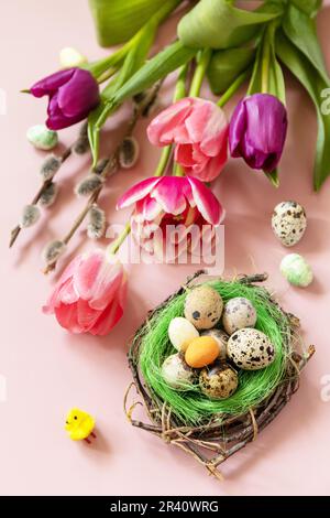 Rosa und lila Tulpen mit bunten Wachteleiern in einem Nest auf rosa Hintergrund. Konzept für Frühlings- und Osterferien. Stockfoto