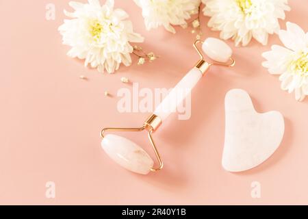 Gesichtsmassagewalze aus Rosenquarz auf pinkfarbenem Hintergrund mit Zigeunerblüten. Massagewerkzeuge mit Jadestein, Anti-Aging, Anti- Stockfoto
