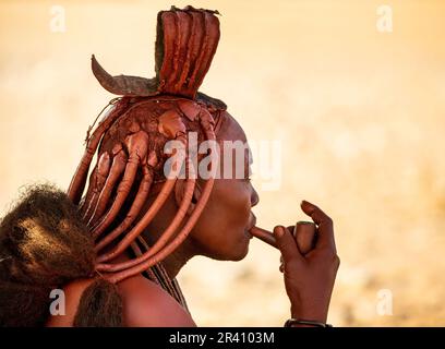 Porträt einer jungen Frau des Himba-Stammes mit einer Raucherpfeife und einer traditionellen Frisur. Stockfoto