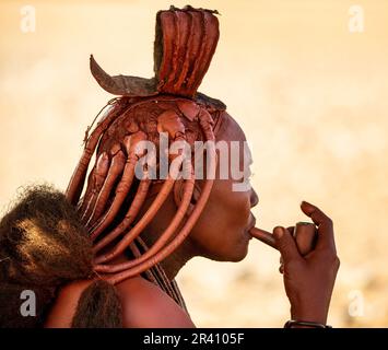 Porträt einer jungen Frau des Himba-Stammes mit einer Raucherpfeife und einer traditionellen Frisur. Stockfoto