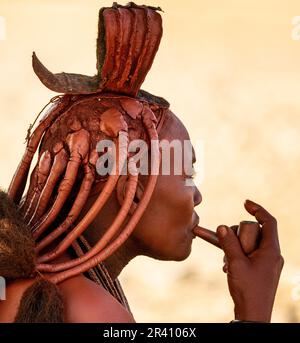 Porträt einer jungen Frau des Himba-Stammes mit einer Raucherpfeife und einer traditionellen Frisur. Stockfoto