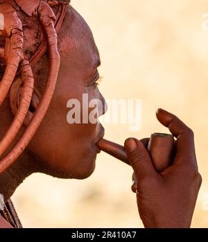 Porträt einer jungen Frau des Himba-Stammes mit einer Raucherpfeife und einer traditionellen Frisur. Stockfoto