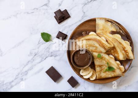 Feier den Pfannkuchen-Tag, gesundes Frühstück. Köstliche hausgemachte Crepes mit Schokoladenkaramell und Bananen auf einem Marmortisch. V Stockfoto