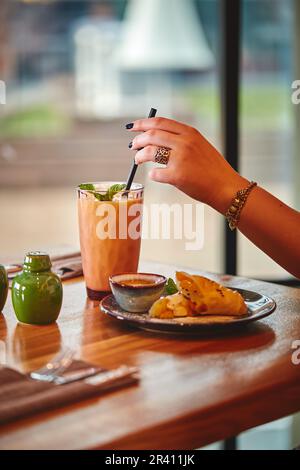 Die Hand einer Frau hält Glas mit Milchshake oder Smoothie auf einem Holztisch im Café vor dem Hintergrund des Fensters Stockfoto