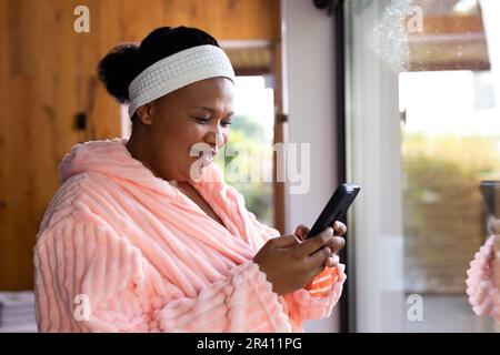 Eine glückliche afroamerikanische Frau in Übergröße, die Bademantel und Kopfband trägt und im Spa ein Smartphone benutzt Stockfoto