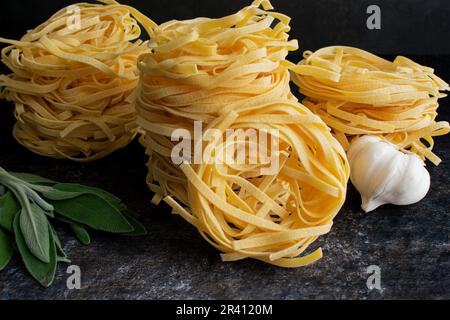 Gestapelte Tagliatelle-Nester mit Salbei und Knoblauch: Nahaufnahme von Seiten und von oben auf getrocknete Nudelnester mit frischen Kräutern Stockfoto